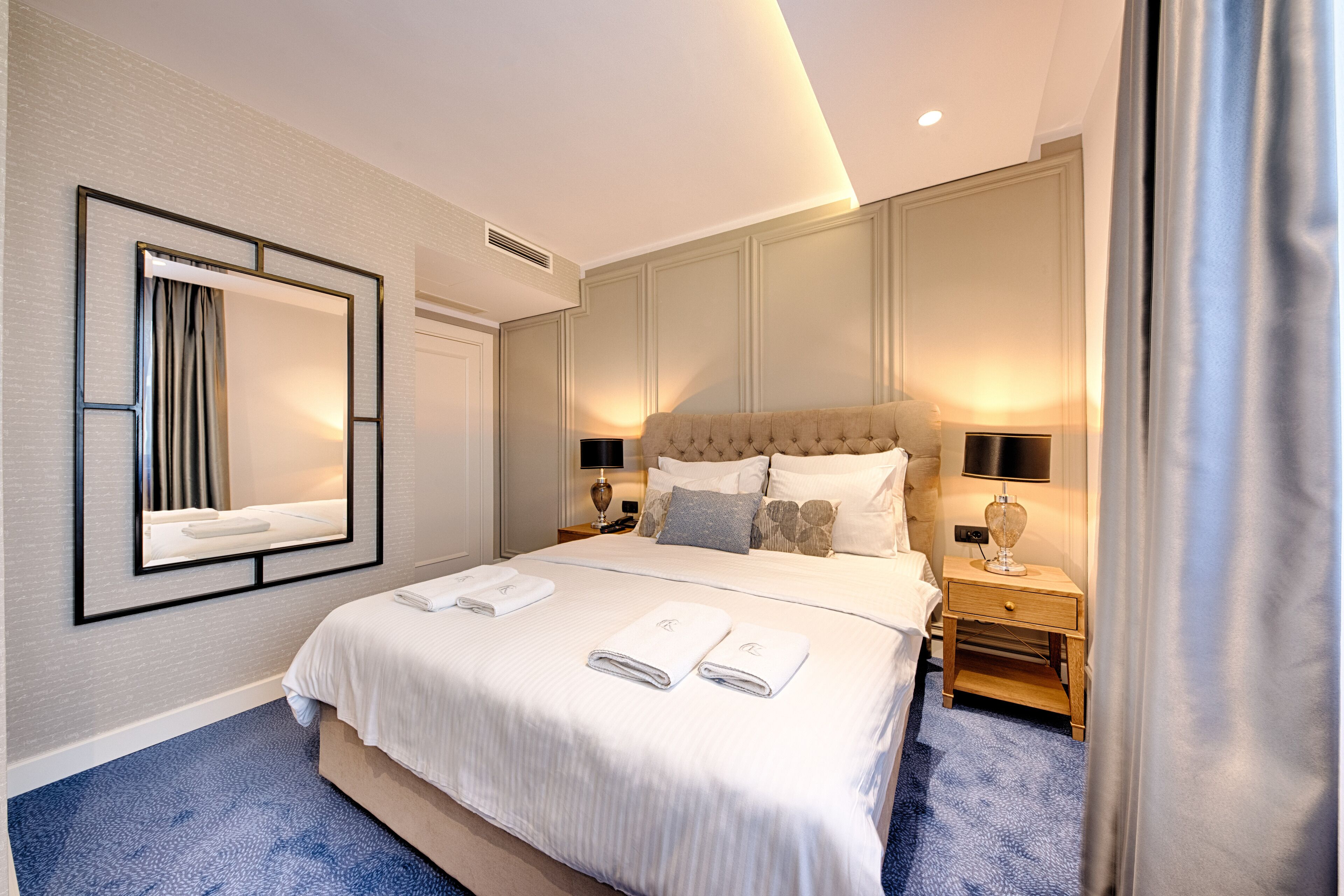 A modern hotel room with a large bed, a mirror, two bedside tables with lamps, and a blue carpet.