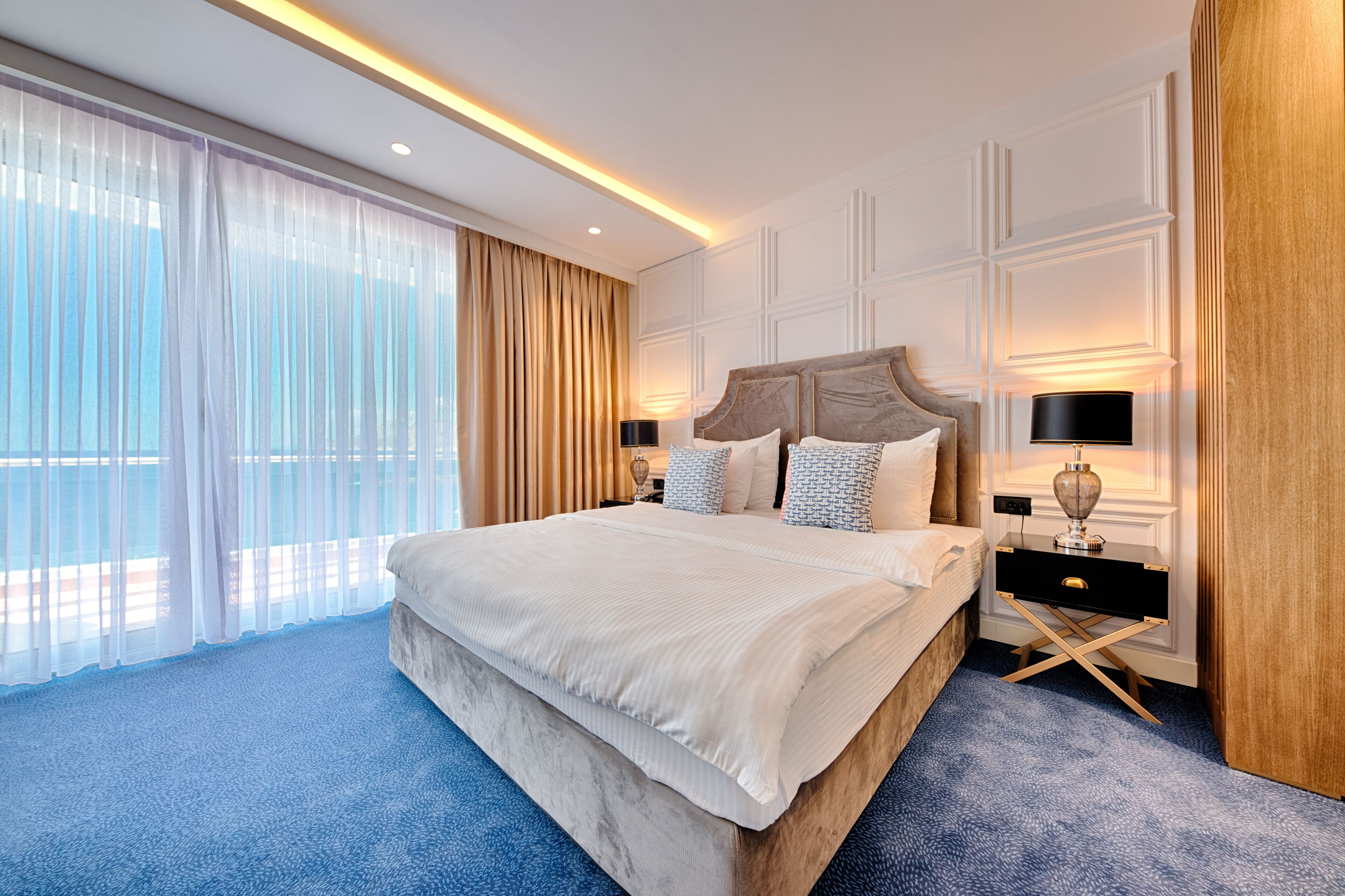 A spacious hotel room with a large bed, a nightstand with a lamp, and a view of the outdoors through sheer curtains.