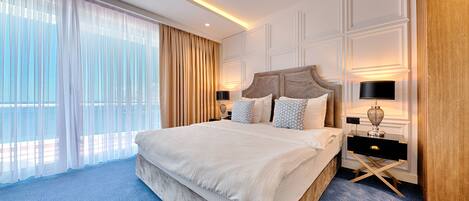 A spacious hotel room with a large bed, a nightstand with a lamp, and a view of the outdoors through sheer curtains.