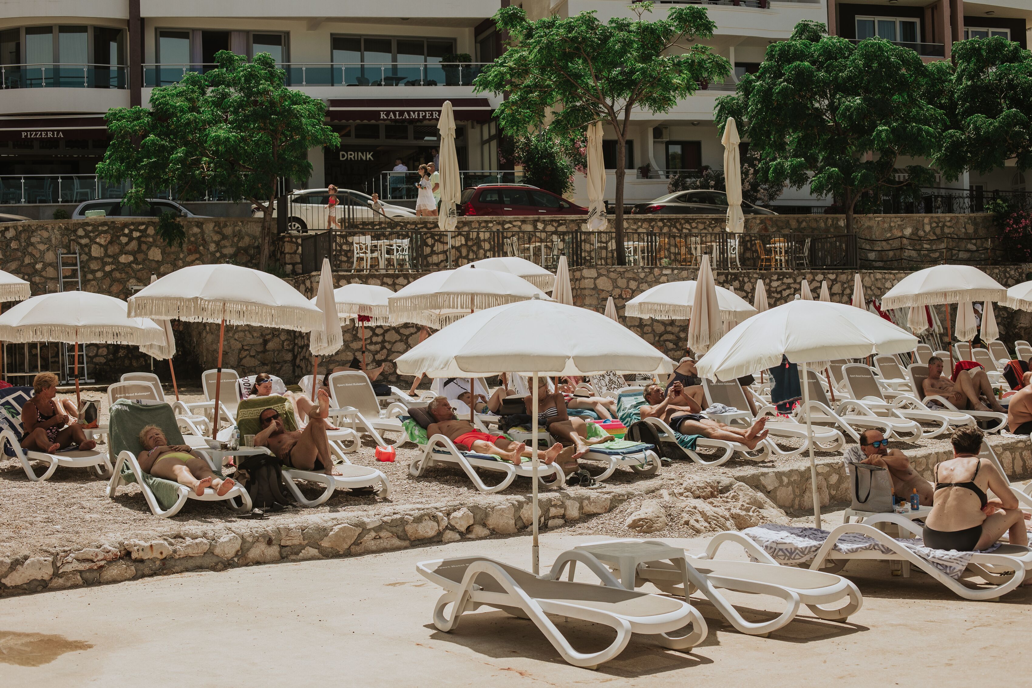 A beachside lounge area with white lounge chairs and umbrellas, a stone wall, and a building with a pizzeria in the background.
