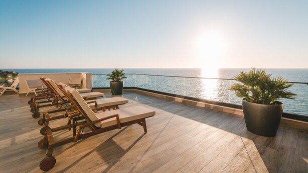 Une terrasse ensoleillée sur le toit, avec des fauteuils en bois et des plantes en pot, d’où l’on peut admirer une mer calme.