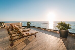 Una terraza en la azotea iluminada por el sol, con sillones de madera y plantas en macetas, con vistas a un mar tranquilo.