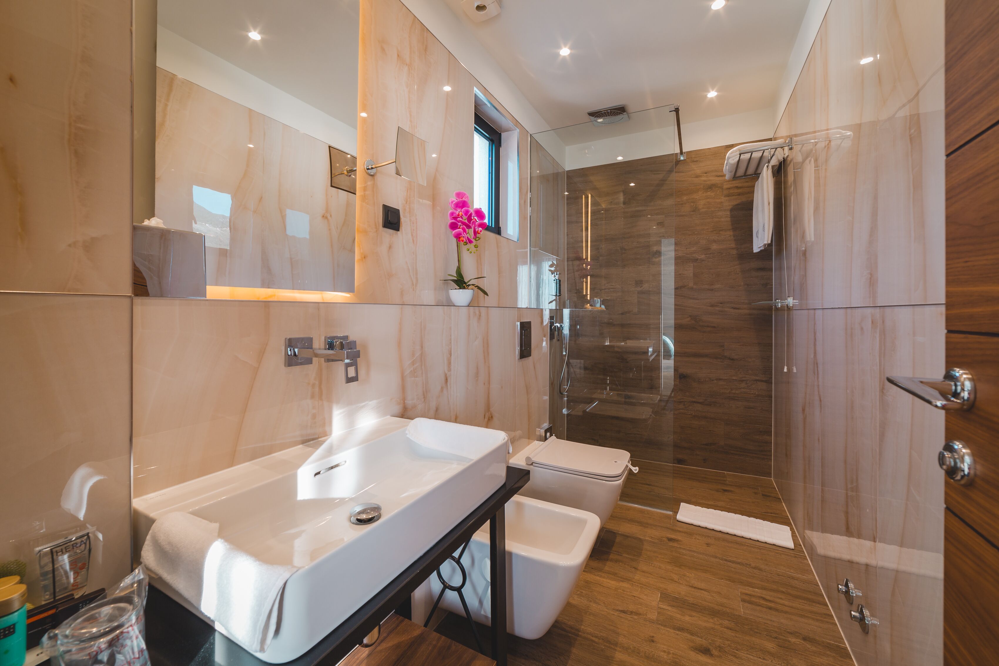 Un baño moderno con un espejo grande, un lavabo blanco, una cabina de ducha de vidrio y suelo de madera.