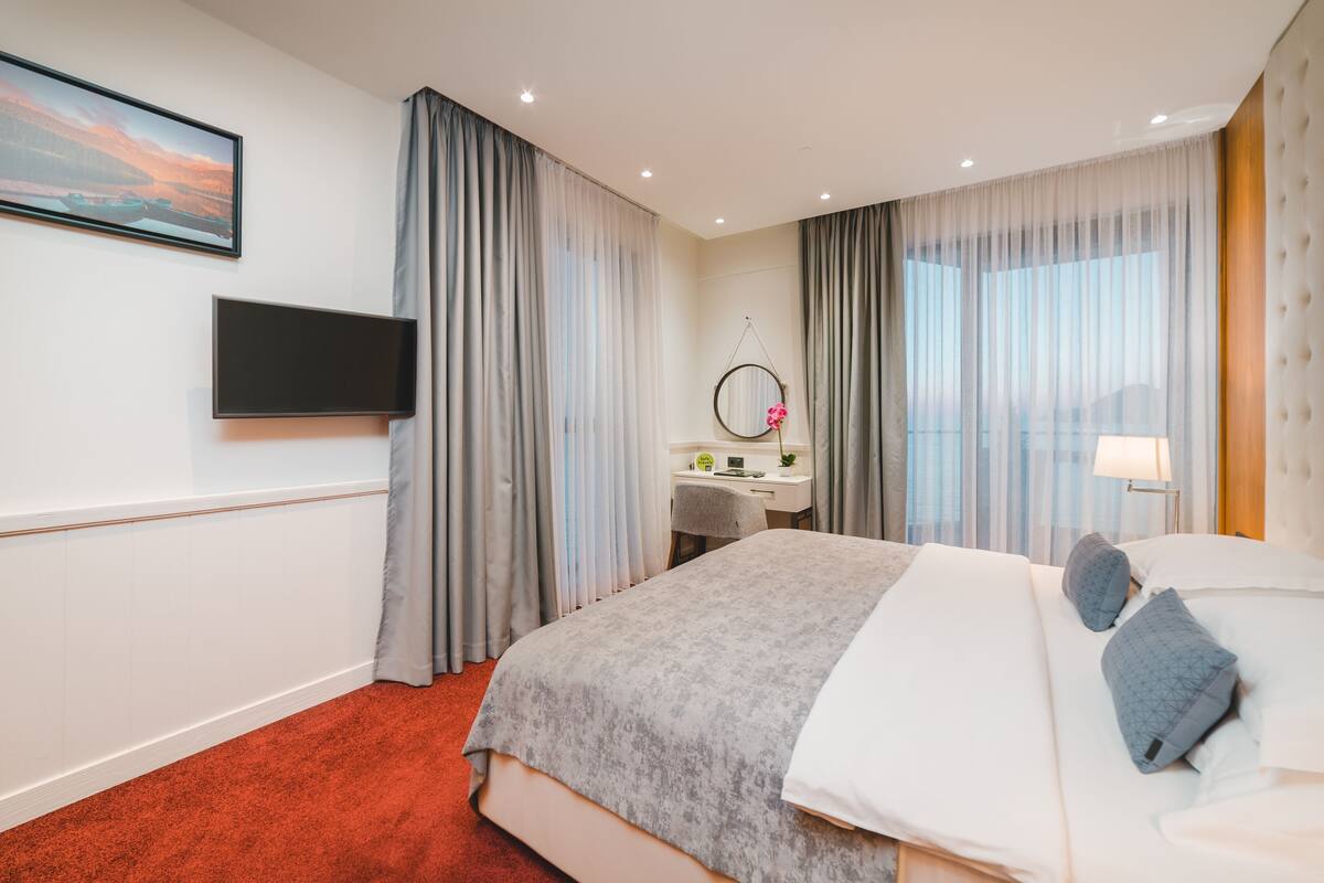 Habitación de hotel con una cama grande, un escritorio con silla, televisor montado en la pared y una ventana con cortinas.