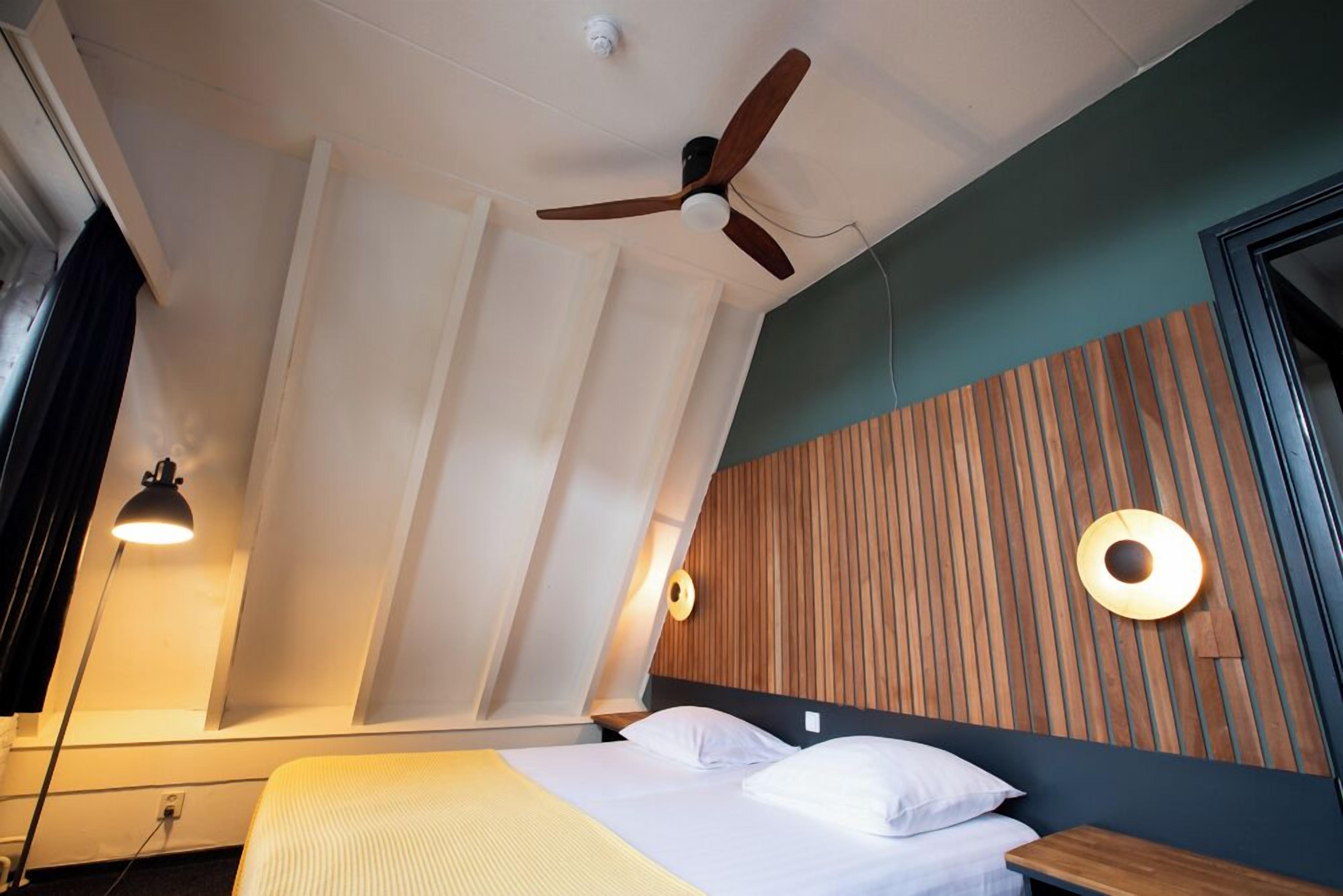 A modern hotel room with a bed, a ceiling fan, a bedside lamp, and a wooden accent wall.