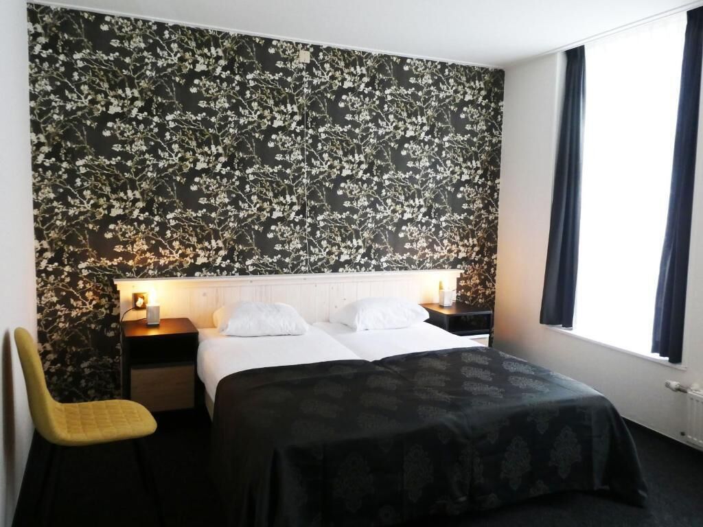 A hotel room with a bed, a nightstand, a chair, and a window with curtains.