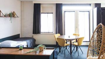 Una habitación con cama, escritorio, silla, mesa y una sillona colgante. Hay ventanas con cortinas, un radiador y una estantería con adornos.