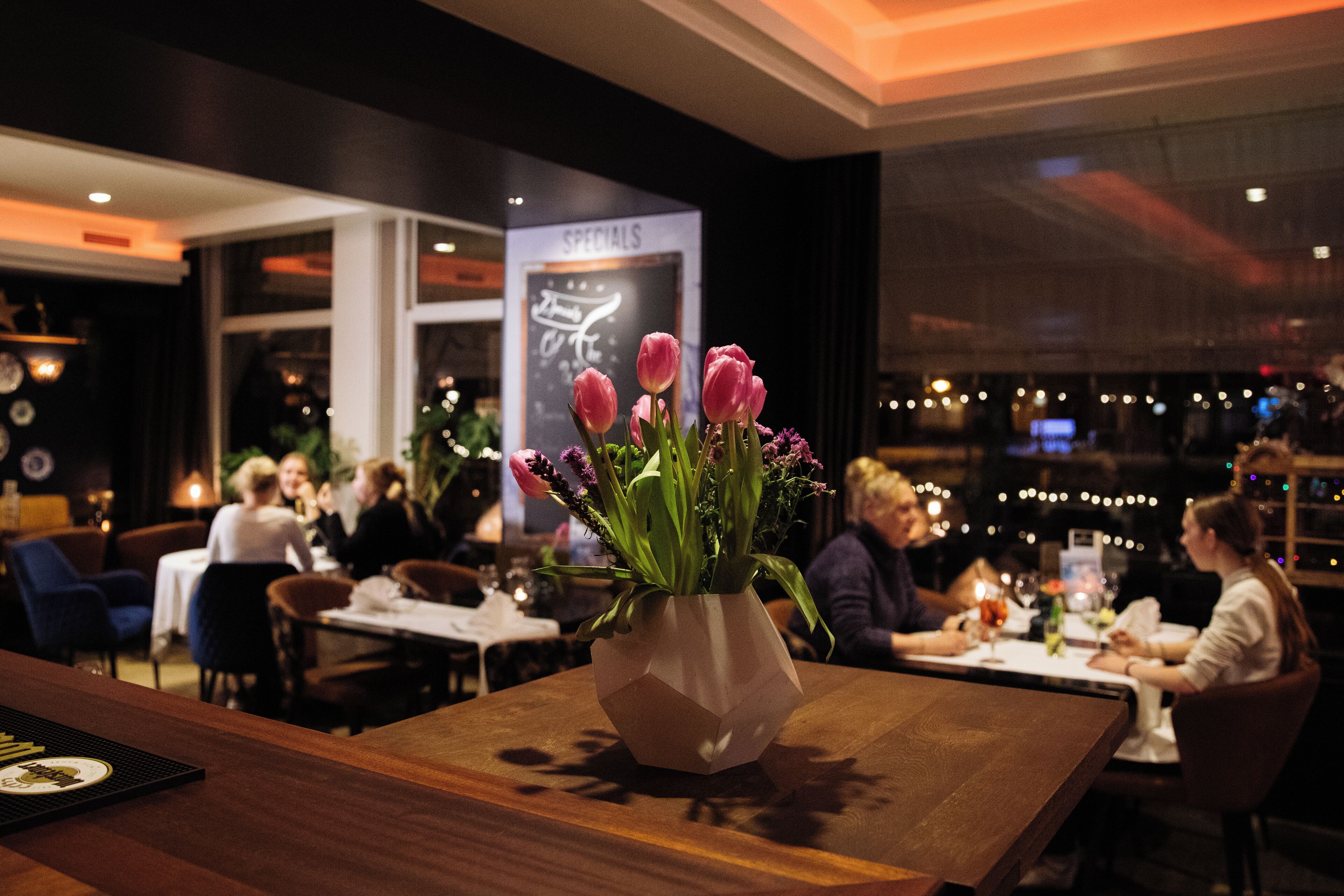 Interior de restaurante con una mesa de madera, un jarrón de flores y comensales al fondo.