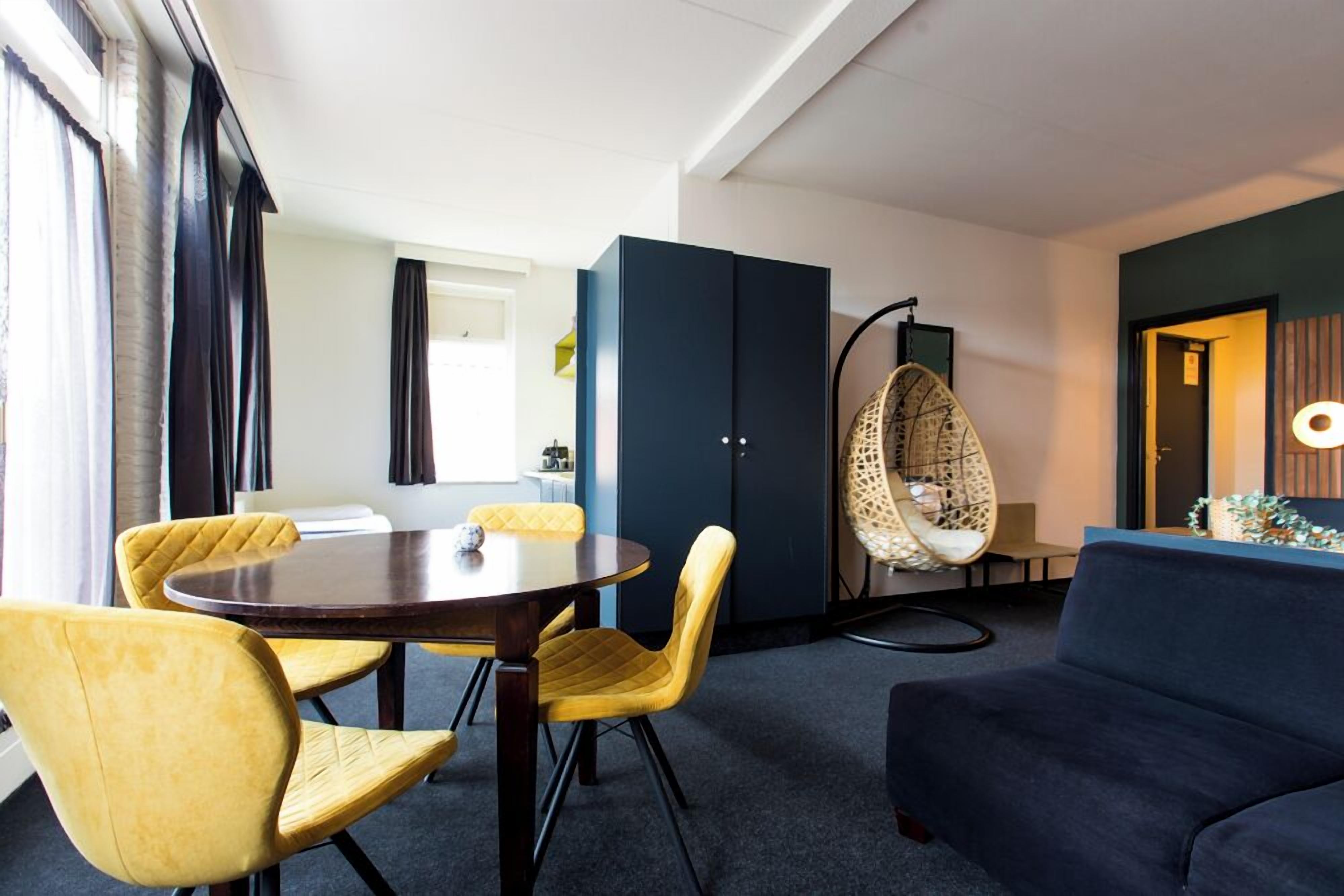 Una sala moderna con un sofá azul oscuro, sillas amarillas, una mesa redonda, un aparador azul oscuro y una silla colgante.