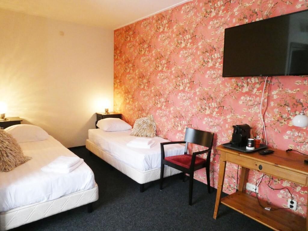 A hotel room with two single beds, a desk with a chair, a television, and a floral wallpaper.