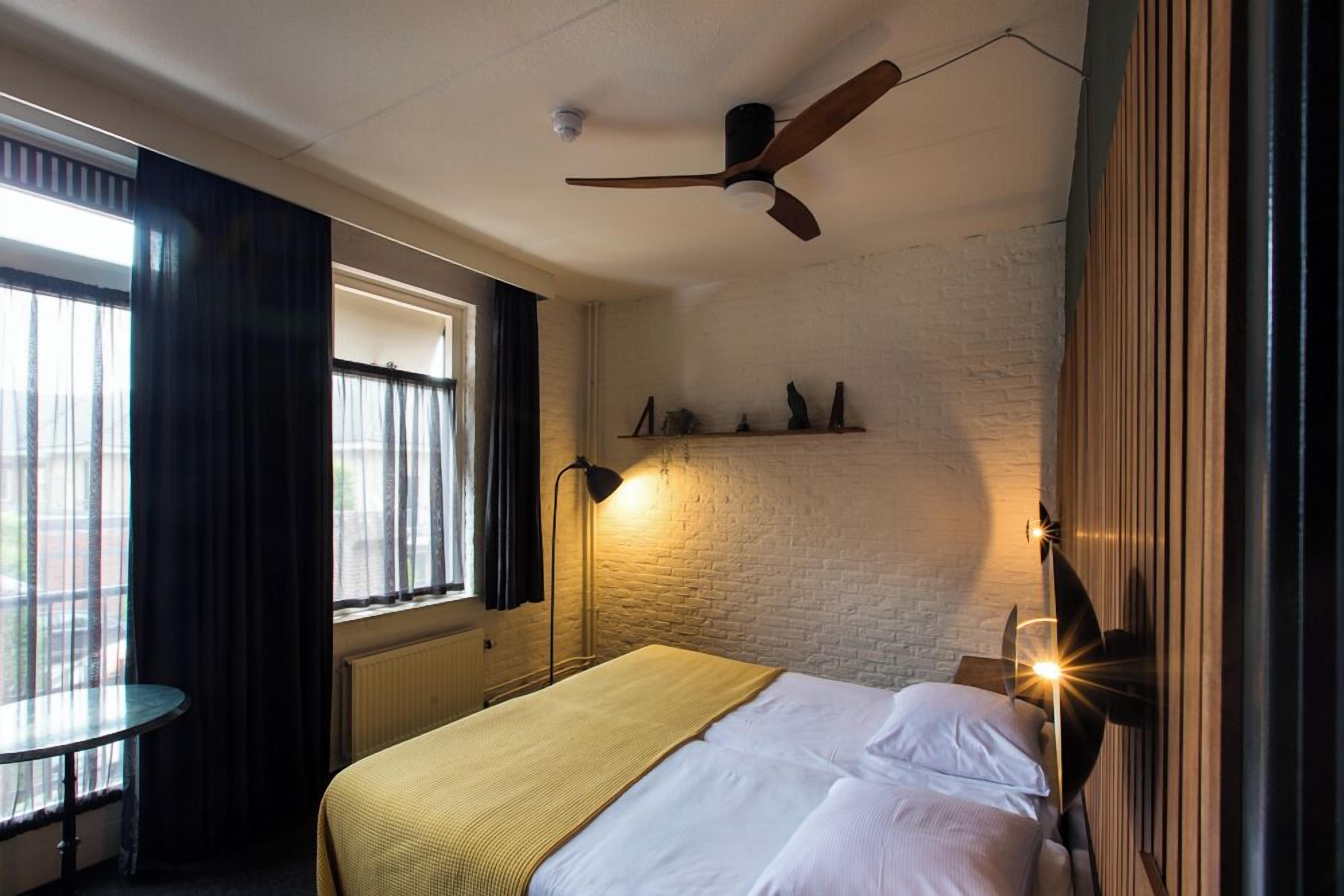 Una habitación de hotel con cama, una mesa redonda, una ventana con cortinas, un ventilador de techo y luces fijadas a la pared.