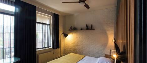 Una habitación de hotel con cama, una mesa redonda, una ventana con cortinas, un ventilador de techo y luces fijadas a la pared.