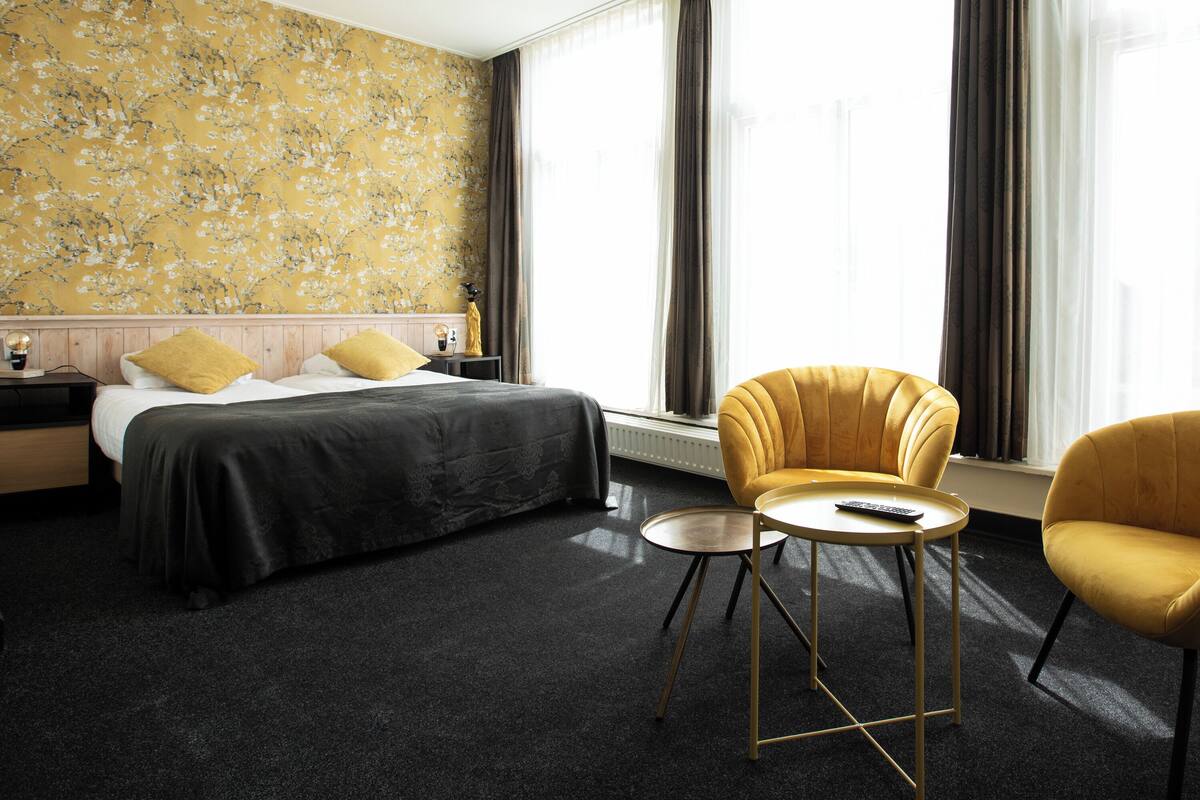 A hotel room with a bed, two yellow armchairs, a small round table, and a large window with curtains.