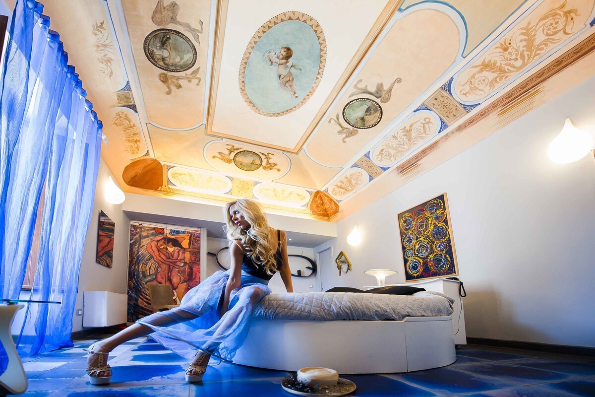 A room with a circular bed, a large painting, and a ceiling with ornate designs.