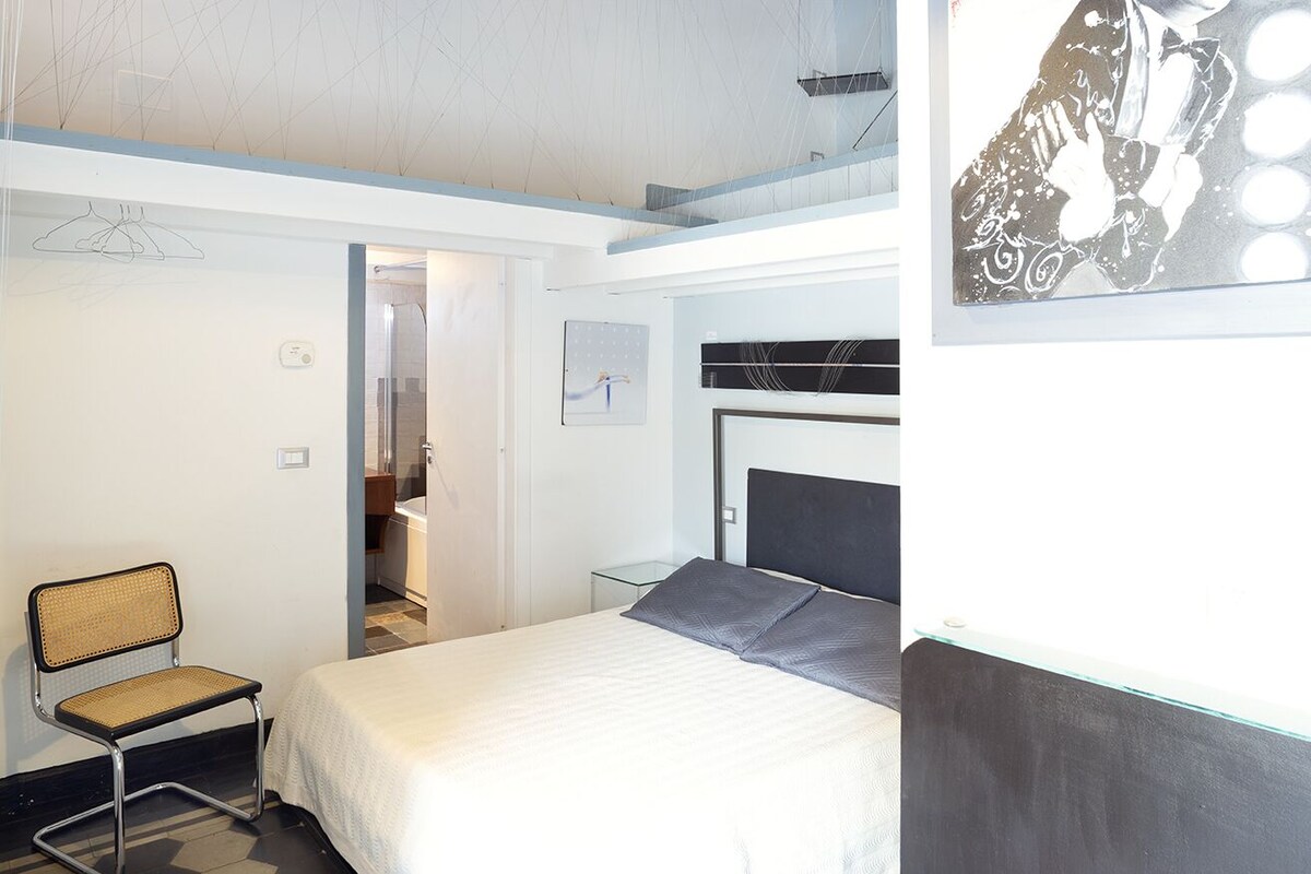 A modern hotel room with a bed, a chair, a small table, and a bathroom visible through an open door.