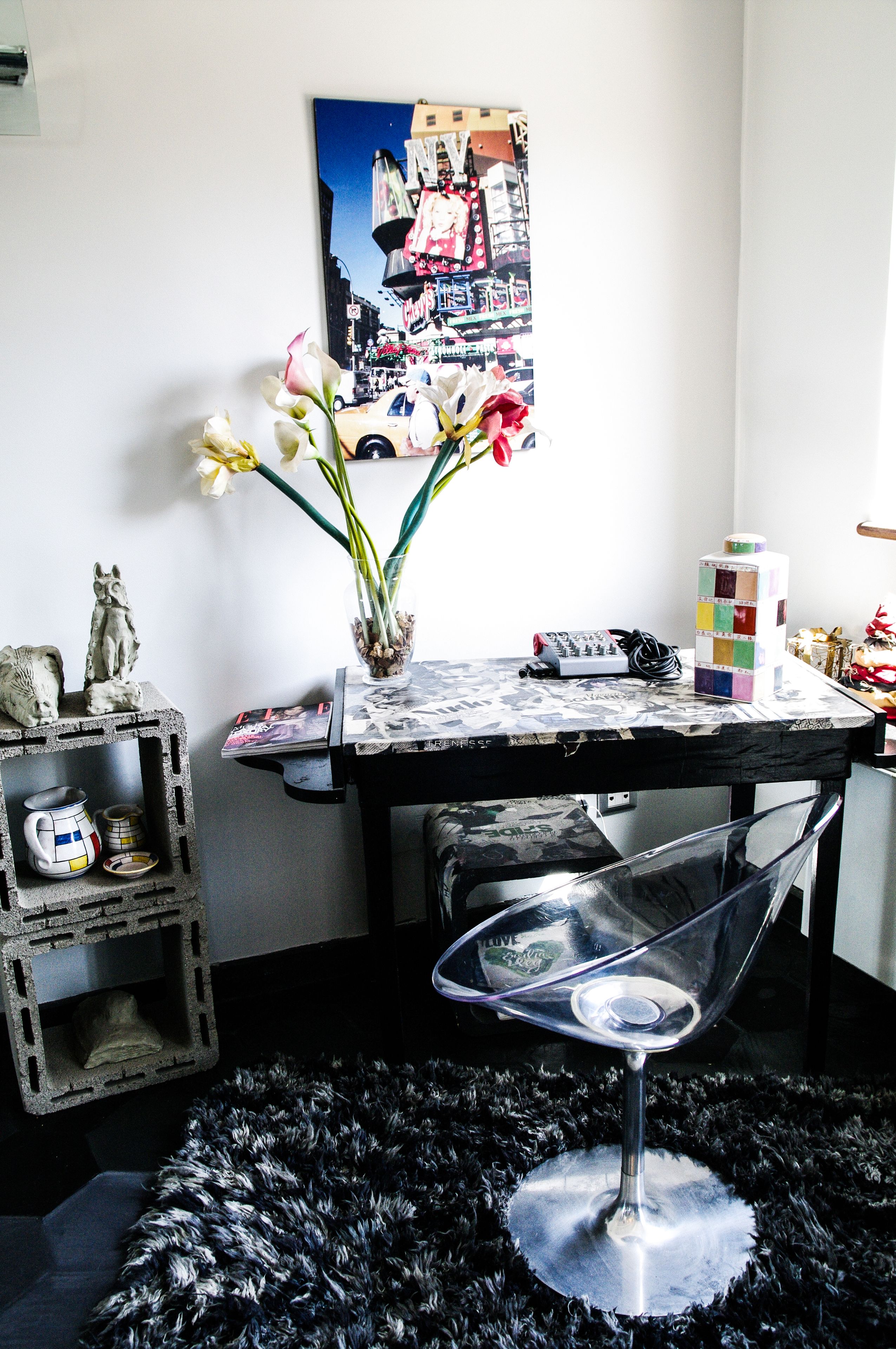 A room with a table, a chair, a vase with flowers, a poster on the wall, and a small shelf with items.