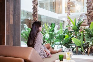 A woman sitting on a beige sofa in a modern indoor space with glass walls, surrounded by lush green plants and a glass table with a green cup and a glass. - The Oasis by Don Carlos Resort (Marbella)