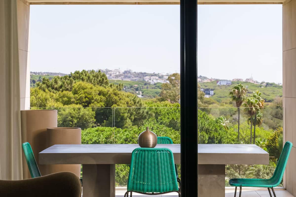 Ein Balkon mit Tisch und Stühlen, der einen Blick auf eine grüne Landschaft und entfernte Gebäude bietet.
