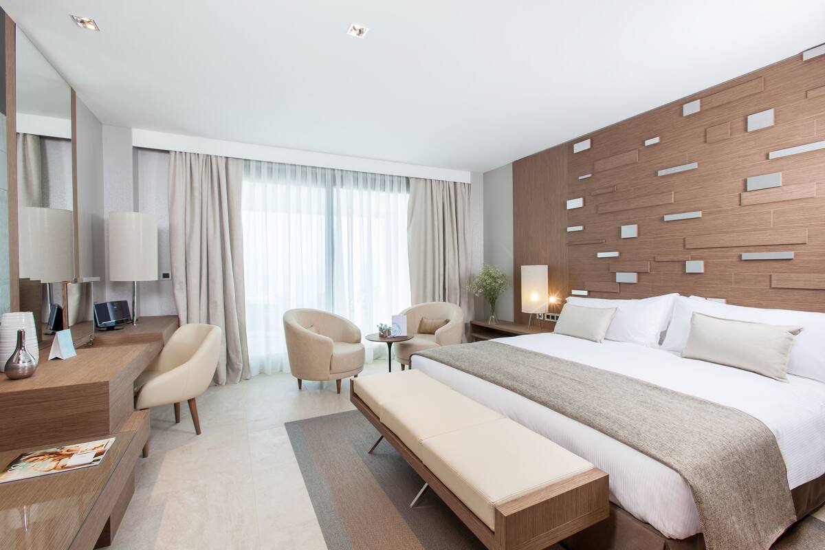 Ein modernes Hotelzimmer mit einem großen Bett, zwei Sesseln, einem Schreibtisch mit Computer und einem kleinen Tisch mit Lampe.