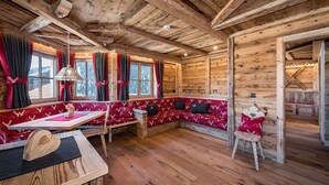 Un interior acogedor de cabaña con techo de madera, una zona de comedor con mesa y bancos, y un rincón de estar con bancos tapizados en rojo.