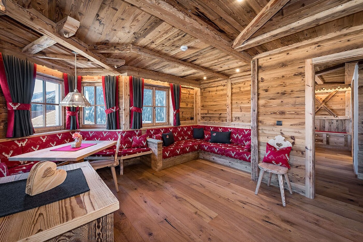 Ein gemütliches Blockhaus-Interieur mit Holzbalkendecke, einem Essbereich mit Tisch und Bänken sowie einer Sitzecke mit rot gepolsterten Bänken.