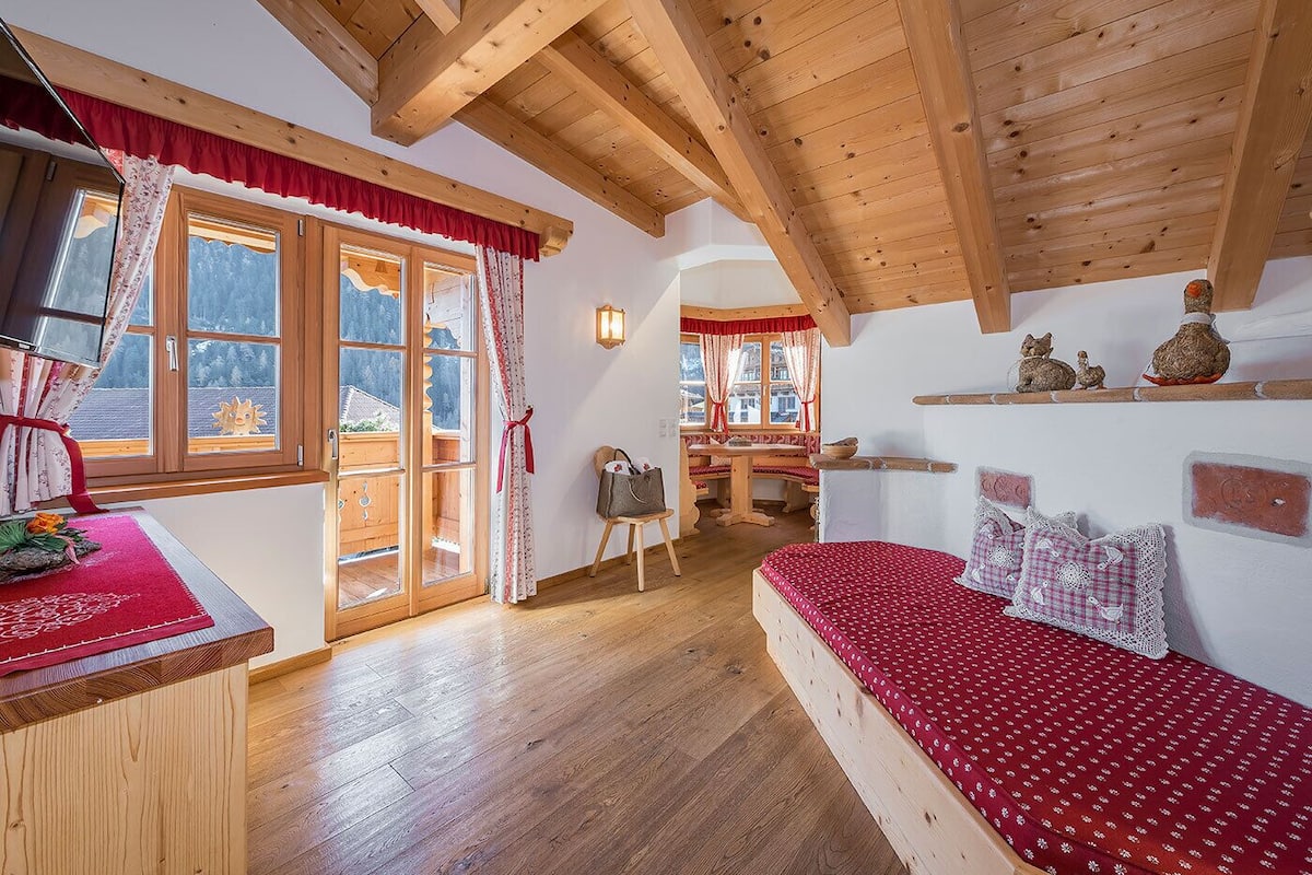 Ein gemütliches Zimmer mit Holzbalken, einem Bett mit roter Bettwäsche, einem Holztisch und Bergblick durch große Fenster.