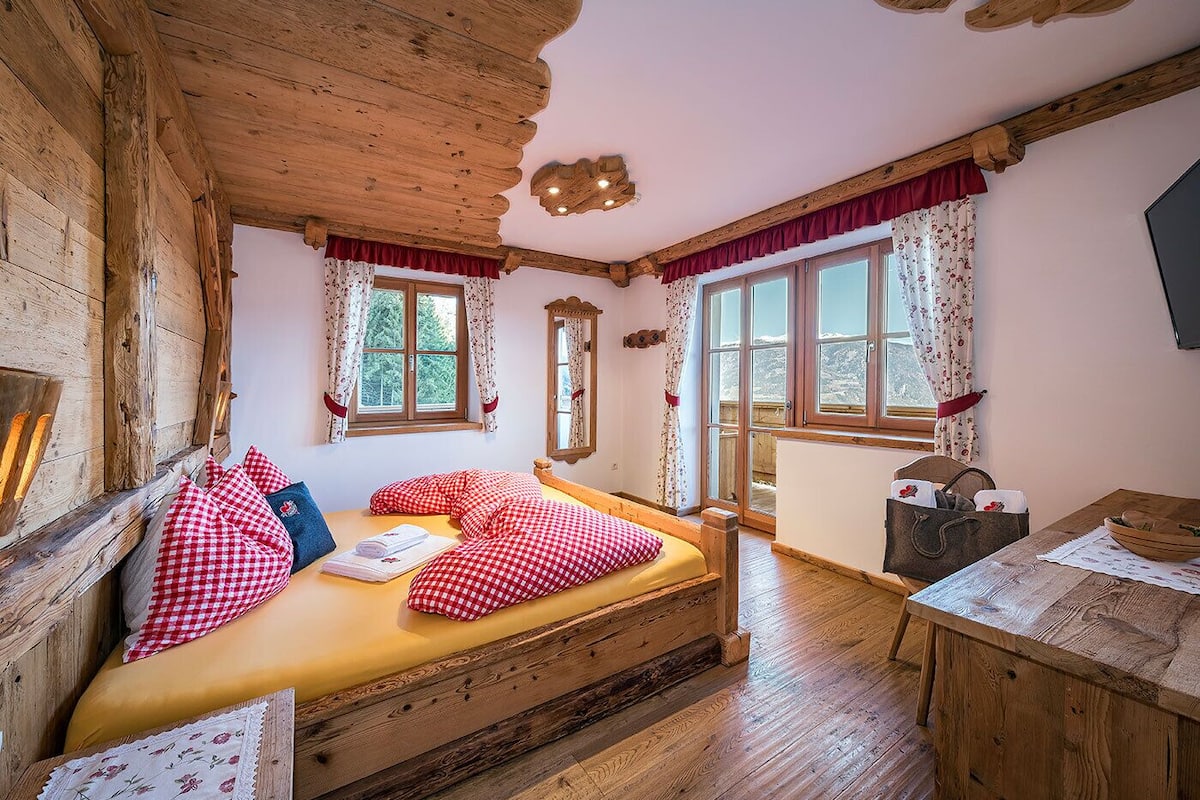 Ein rustikaler Holzraum mit einem Bett, zwei mit Vorhängen versehenen Fenstern, einem Holztisch und einem Kamin.
