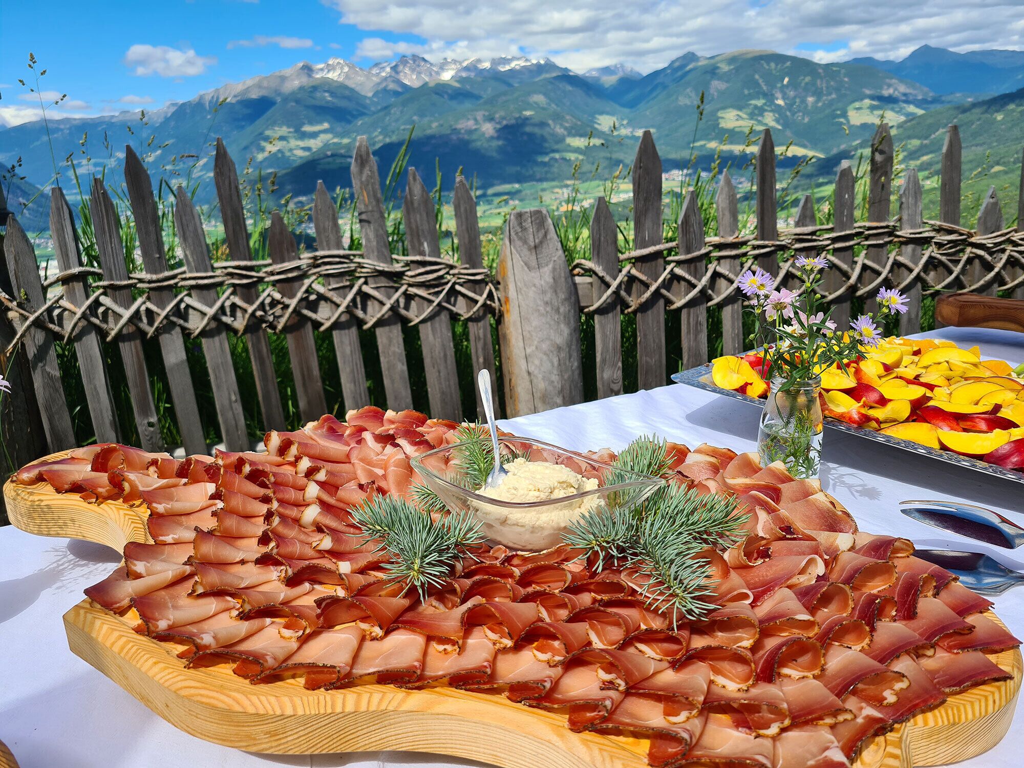 Una tabla de fiambres con jamón serrano y un cuenco de salsa, con montañas al fondo.
