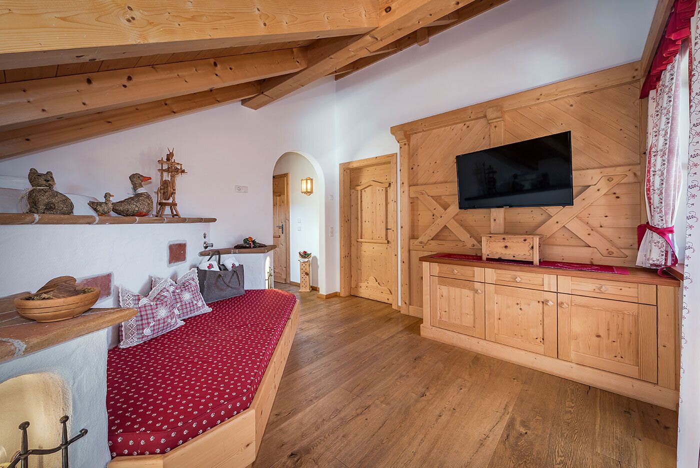 A cozy room with a wooden ceiling, a bed with a red patterned bedspread, a wooden cabinet with a flat-screen TV, and rustic decor including wooden wall shelves and a small table with a lamp.