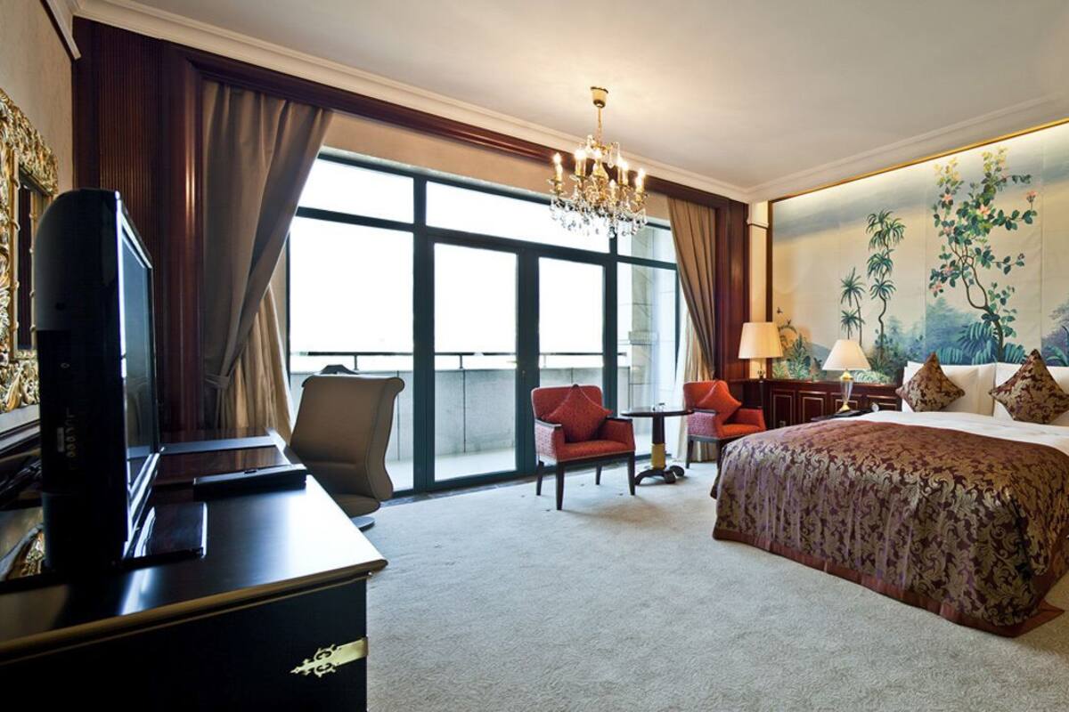 A hotel room with a large bed, a desk, a TV, and a painting of tropical plants on the wall.