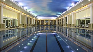 Un'ampia piscina coperta con soffitto a specchio, circondata da poltrone relax e un'imponente architettura.