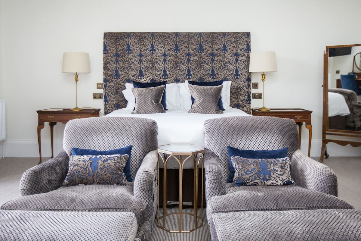 A hotel room with a large bed, two armchairs, a small table, a mirror, and a patterned headboard.