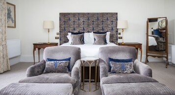 A hotel room with a large bed, two armchairs, a small table, a mirror, and a patterned headboard.