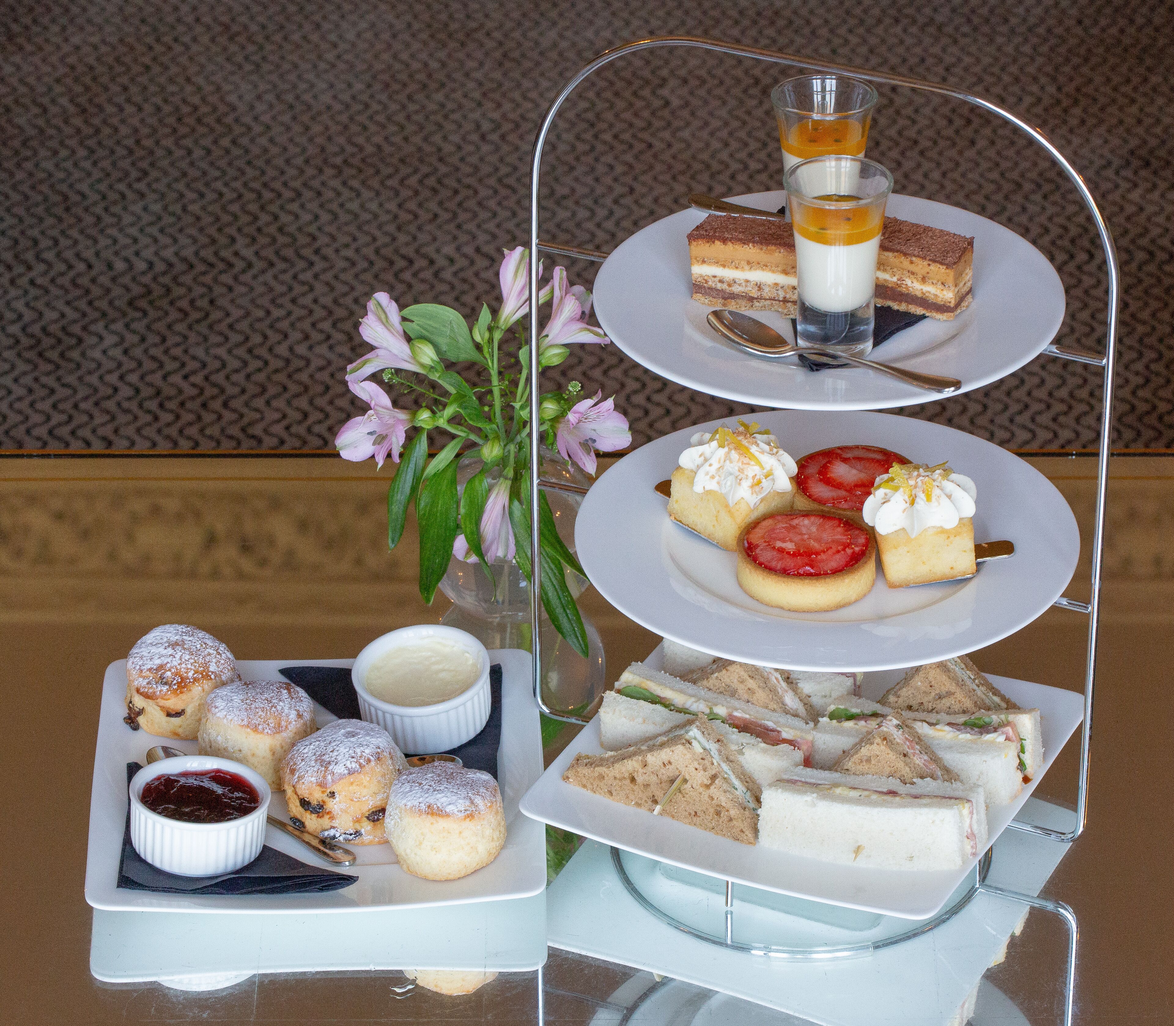A three-tiered dessert stand with various pastries and desserts, including scones, sandwiches, and a small vase of flowers.