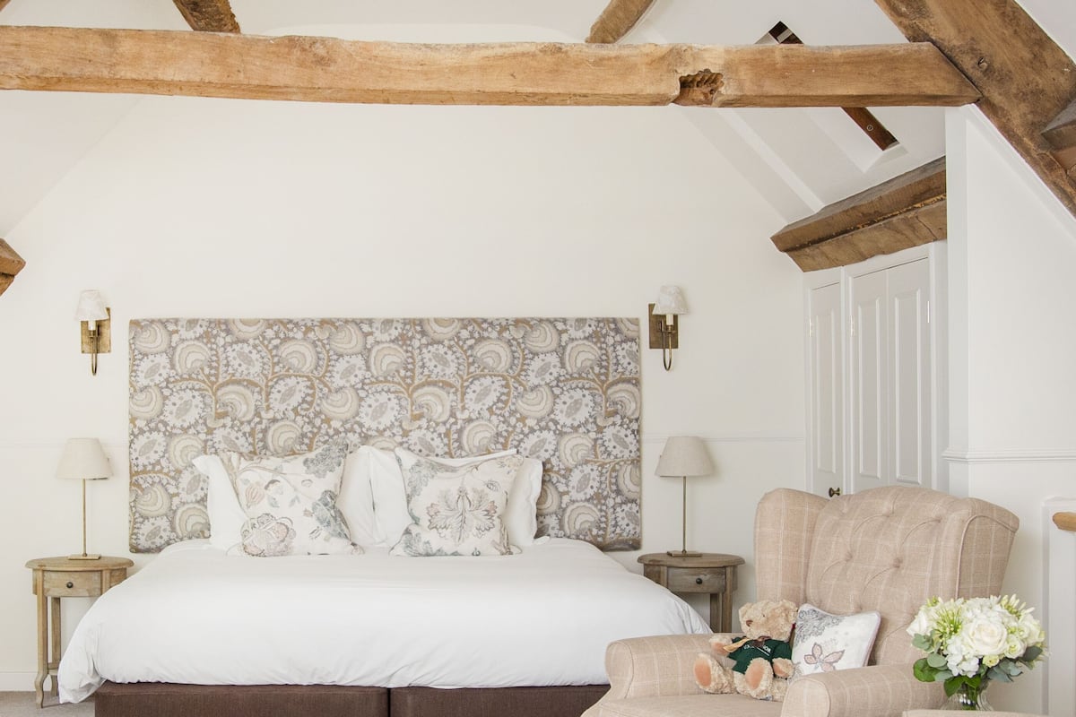 A bedroom with a large bed, wooden beams, a headboard with a floral pattern, bedside tables with lamps, and a chair with a teddy bear.