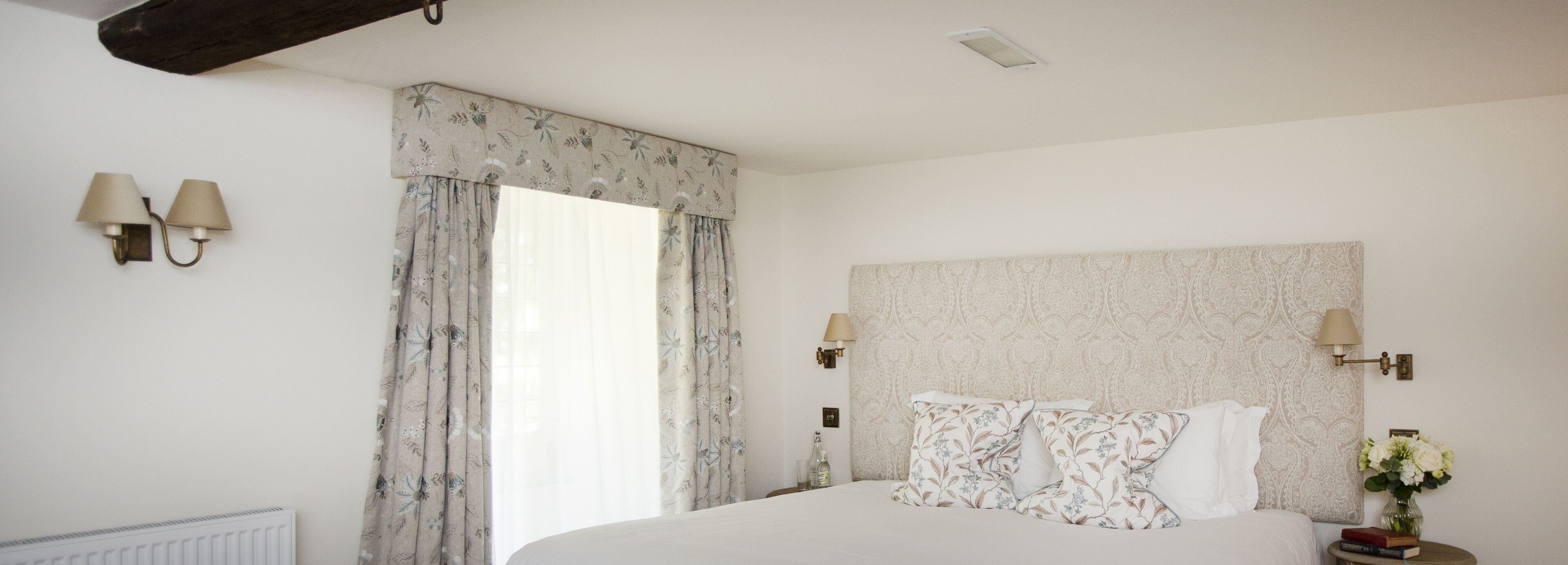 Une chambre à coucher avec un grand lit, des rideaux à motifs floraux, deux lampes fixées au mur et une tête de lit décorée.