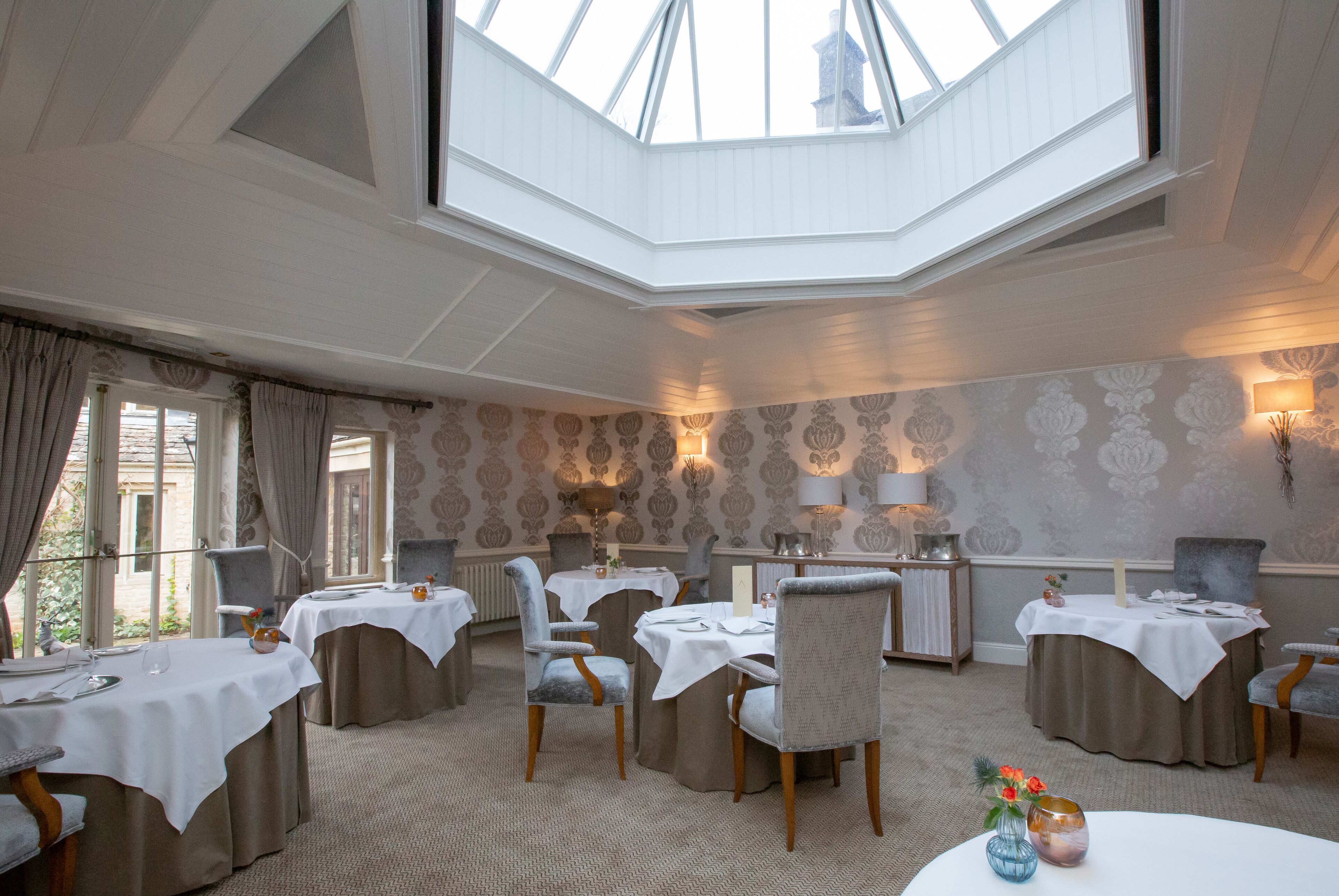 A dining area with tables set for four, a high ceiling with a skylight, and patterned wallpaper.