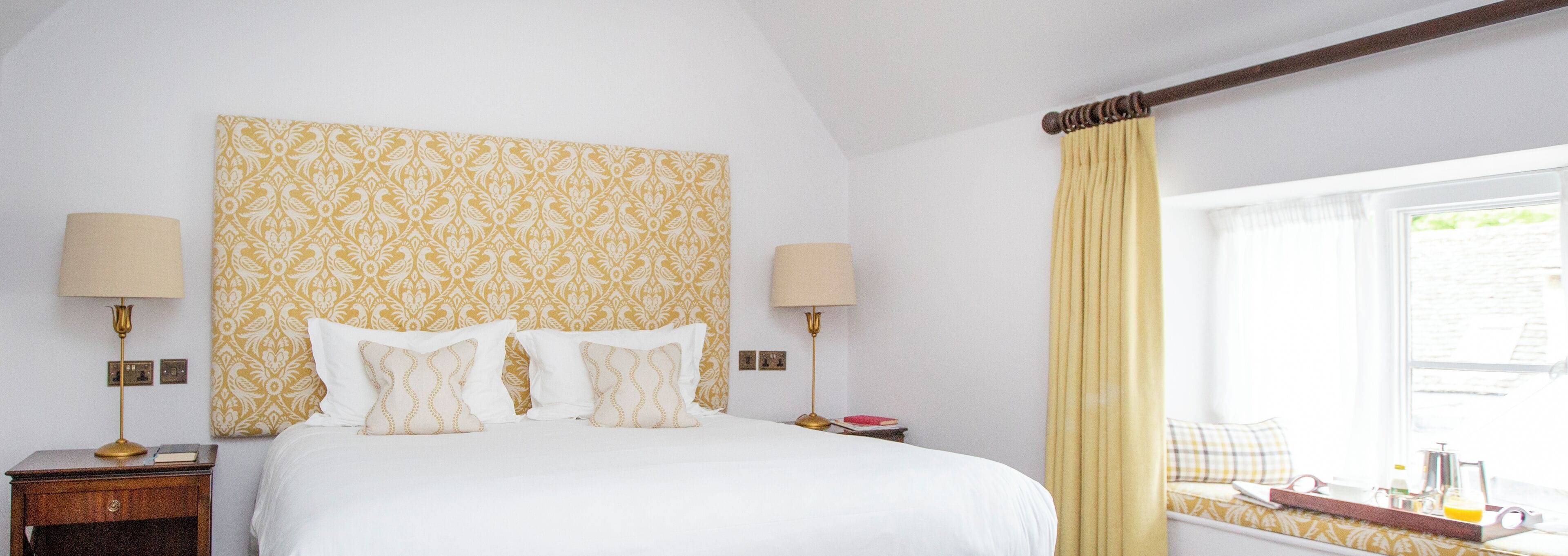 A hotel room with a bed, two bedside lamps, a window with curtains, and a bedside table with a lamp and a book.