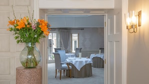 Un comedor con mesas preparadas para dos, un florero sobre un pedestal y vista al salón a través de una puerta abierta.