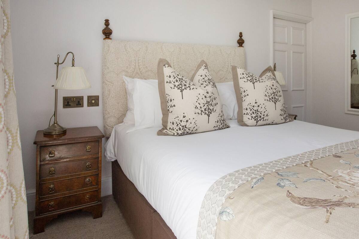 A bedroom with a bed, a nightstand with a lamp, a dresser, and a window with a mirror.