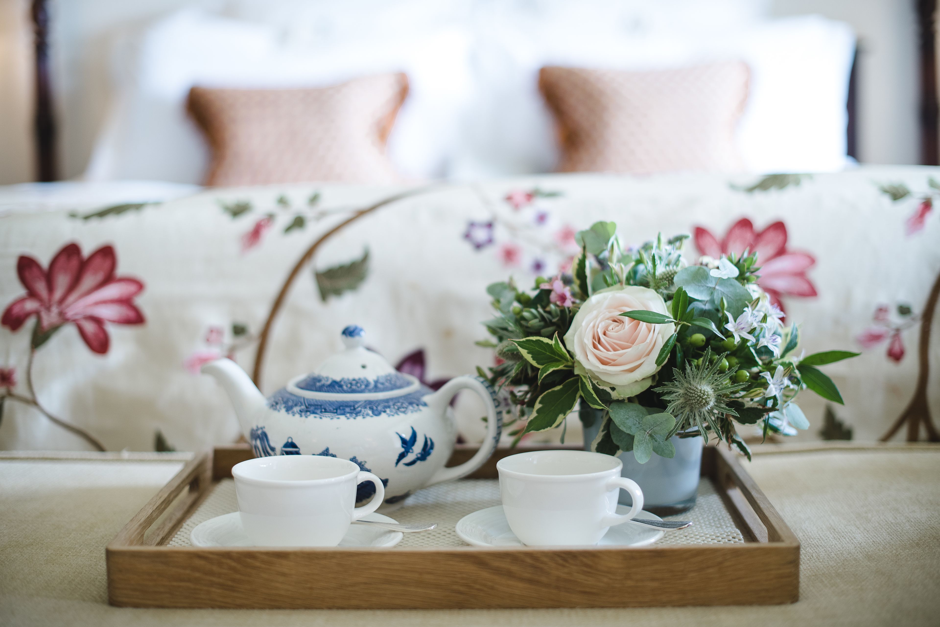 A tray with a teapot, two cups, and a floral arrangement on a bed with a floral bedspread.