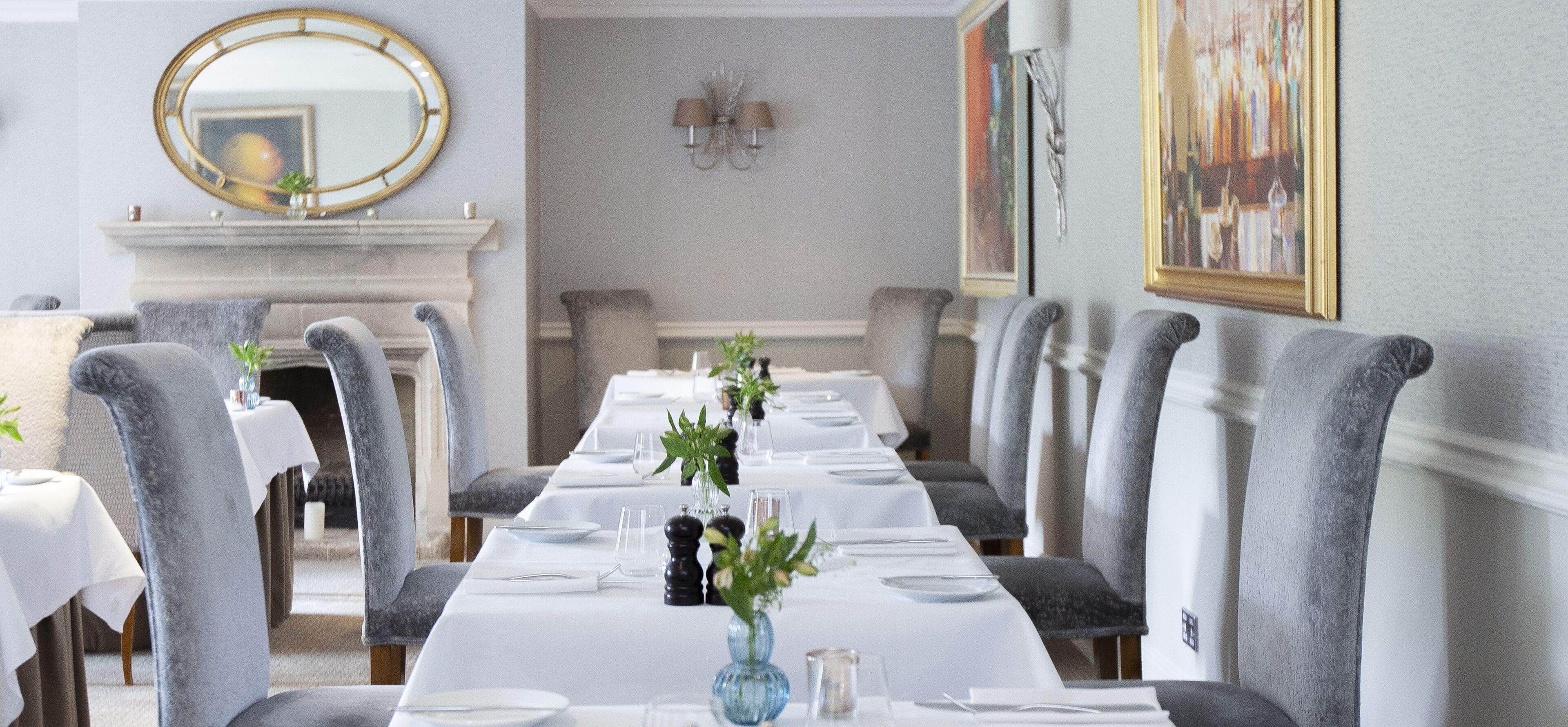 A formal dining room with a long table set for a meal, grey upholstered chairs, a fireplace, and a chandelier.