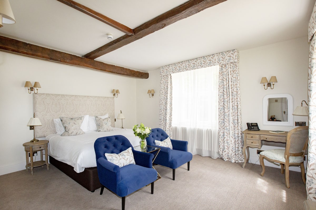 A hotel room with a large bed, two blue armchairs, a desk with a mirror, and a window with floral curtains.