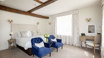 A hotel room with a large bed, two blue armchairs, a desk with a mirror, and a window with floral curtains.