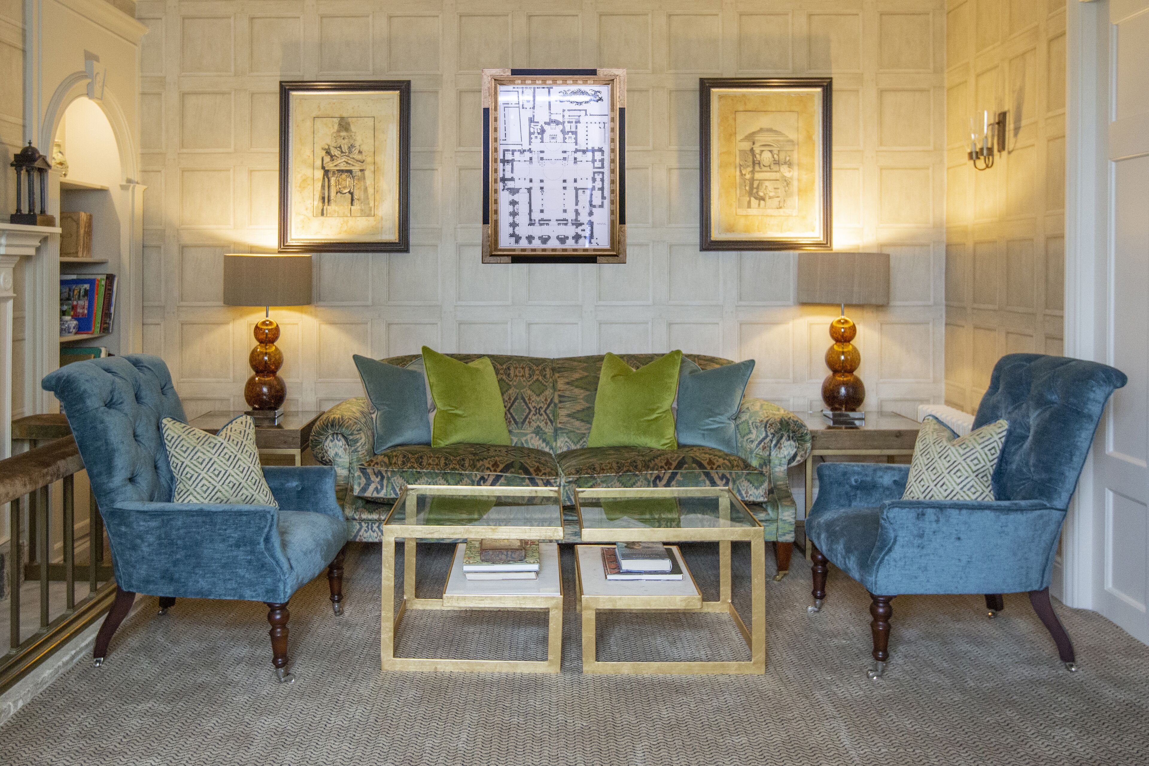 A room with a patterned sofa, two armchairs, and a glass coffee table. There are framed artworks on the walls and a fireplace.