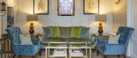 A room with a patterned sofa, two armchairs, and a glass coffee table. There are framed artworks on the walls and a fireplace.