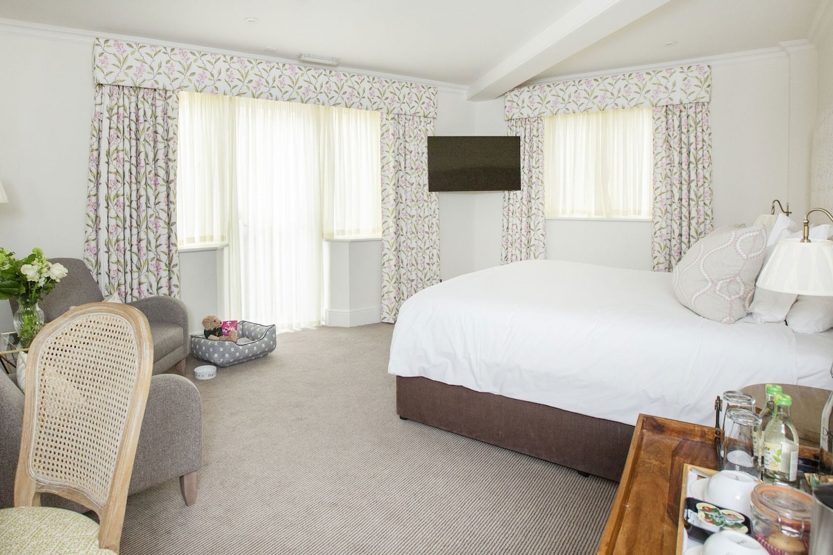 Habitación de hotel con cama, una silla, mesita de noche, lámpara, televisor y cortinas con estampado floral.