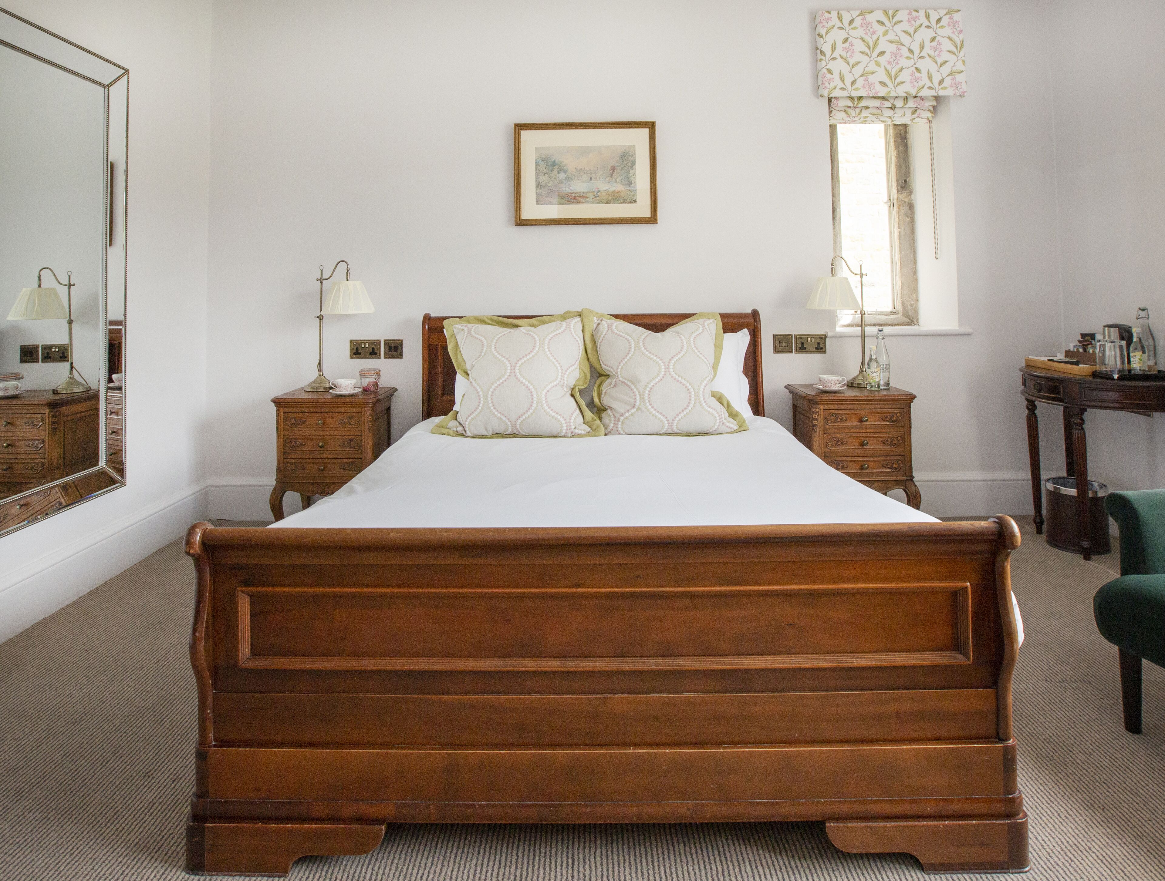 A neatly arranged bedroom with a large wooden bed, two bedside tables with lamps, a framed picture on the wall, and a window with floral curtains.