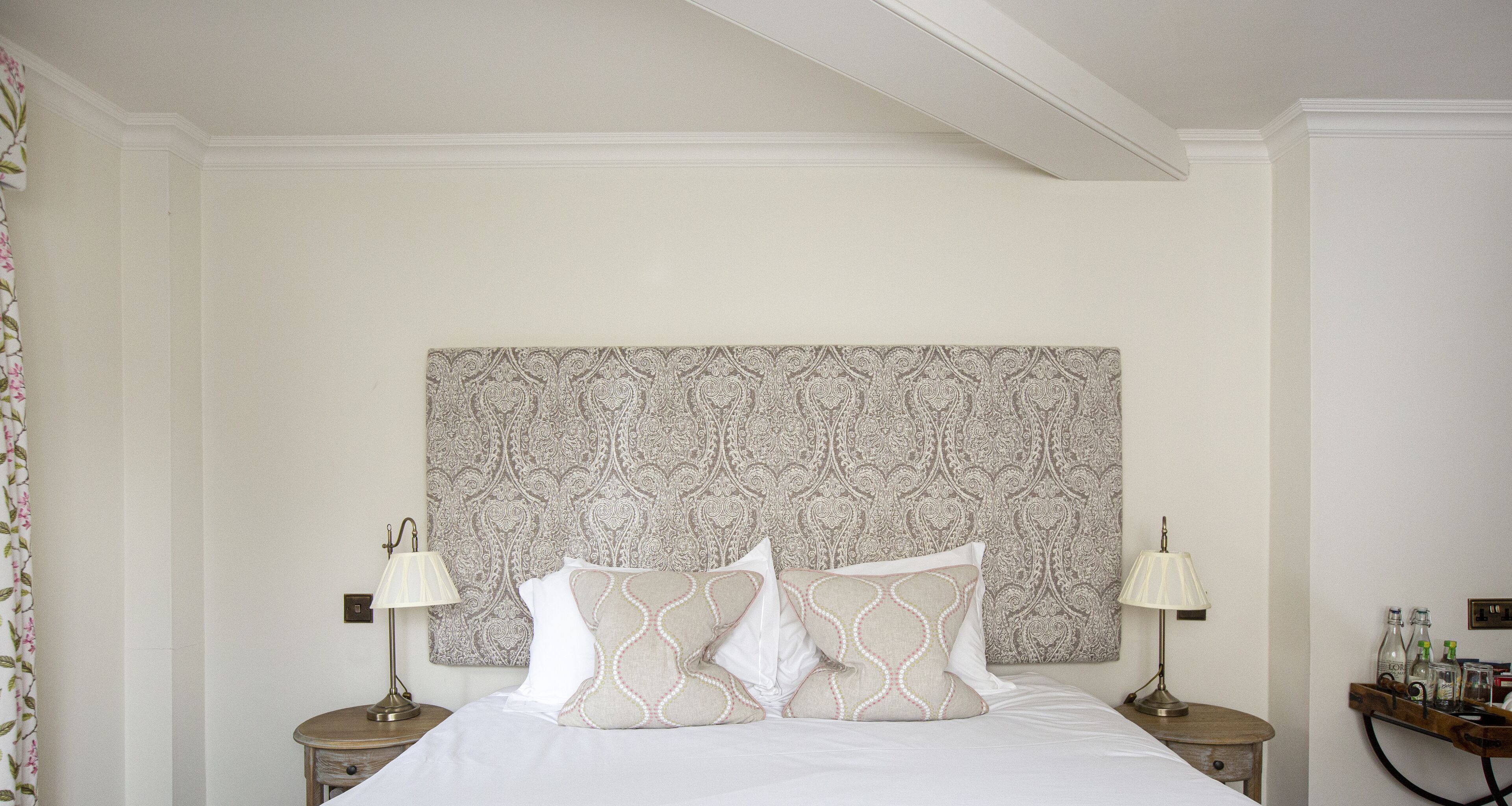 A neatly arranged hotel room with a large bed, two bedside tables with lamps, a patterned headboard, and a small side table with bottles.