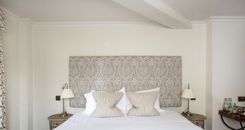 Habitación de hotel ordenada con una cama grande, dos mesitas de noche con lámparas, un cabecero estampado y una pequeña mesita con botellas.